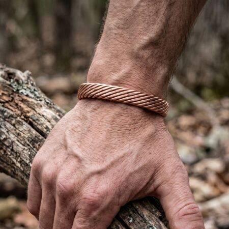 A close-up of a man's arm wearing a tightly woven pure copper cuff bracelet, with his hand resting on a rugged tree branch outdoors