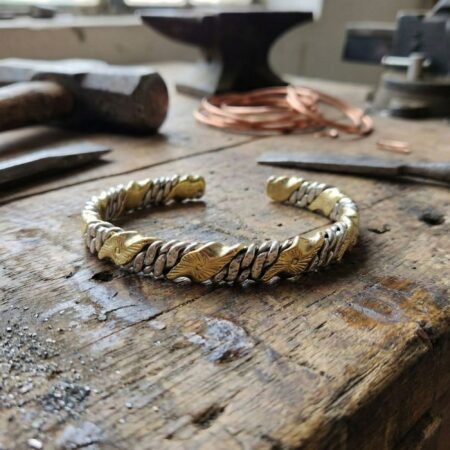 A high-angle shot of the twisted brass and steel bracelet resting on a scarred wooden workbench. In the softly blurred background, there are blacksmithing tools including a hammer, a chisel, and coils of copper wire, evoking a handcrafted metalwork atmosphere.