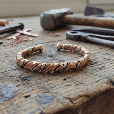 A finished twisted wire cuff bracelet centered on a jeweler's workbench, with chisels, metal files, and raw metal cuttings in the background.