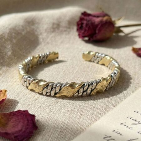 The mixed-metal cuff bracelet placed gently on wrinkled natural beige linen. It is accompanied by scattered dried red rose petals and a blurred handwritten paper note, creating a soft, warm, and artisanal mood.