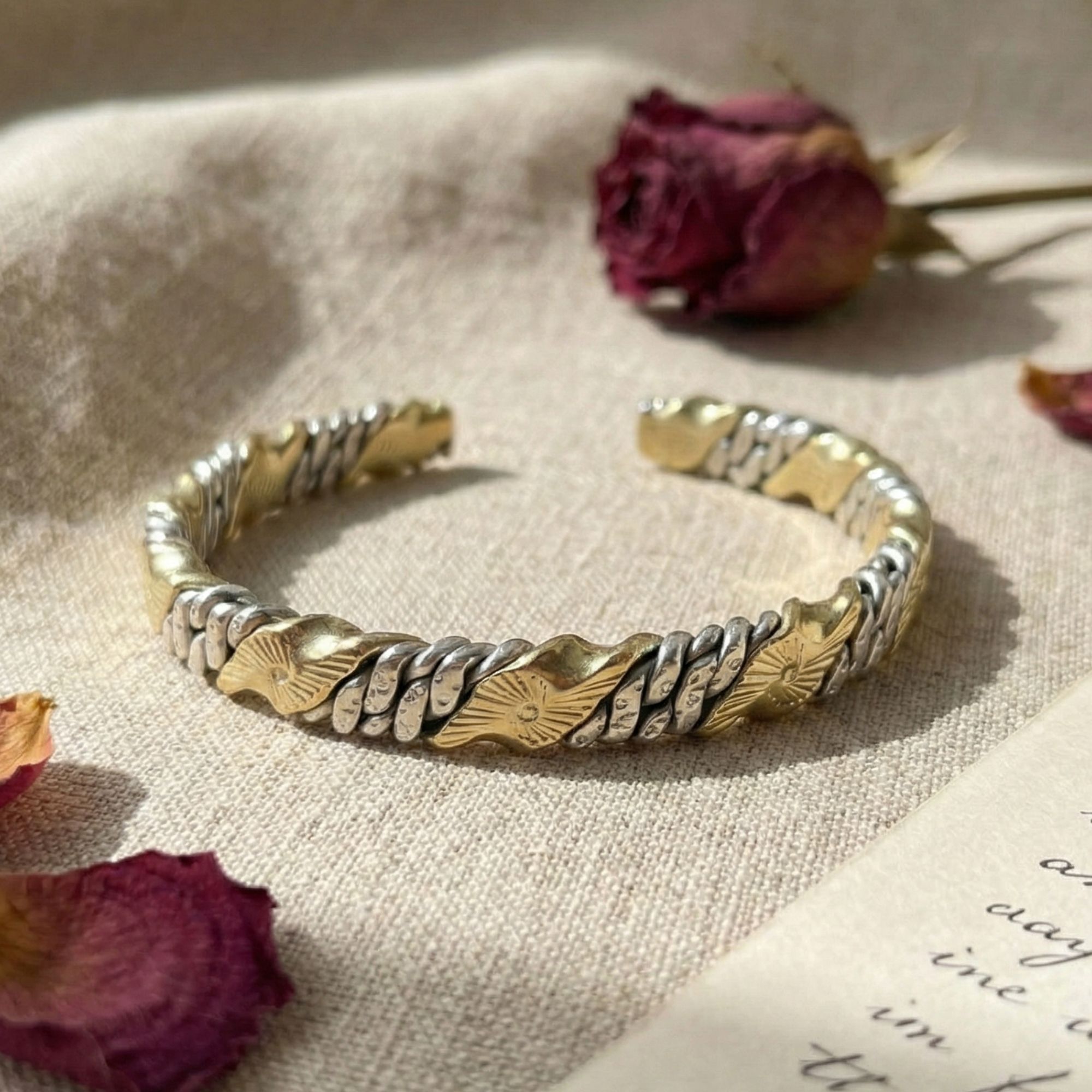 The mixed-metal cuff bracelet placed gently on wrinkled natural beige linen. It is accompanied by scattered dried red rose petals and a blurred handwritten paper note, creating a soft, warm, and artisanal mood.