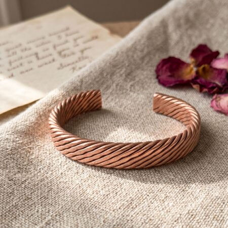 A rustic woven copper cuff resting on natural linen fabric, styled next to a handwritten vintage letter and dried red rose petals
