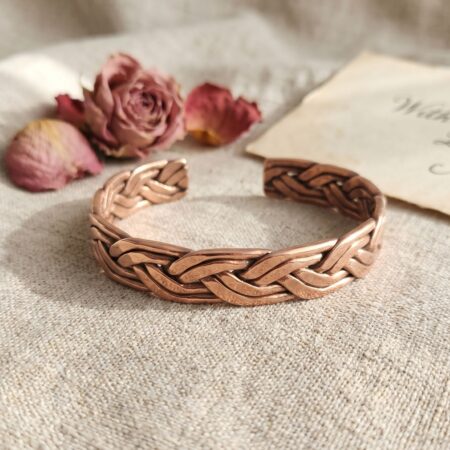 A close-up photograph of a heavy, handcrafted men's cuff bracelet made of tightly braided pure copper wires. The bracelet rests on a dark, textured charcoal gray slate background under bright light, highlighting the raw metal texture and intricate weave pattern