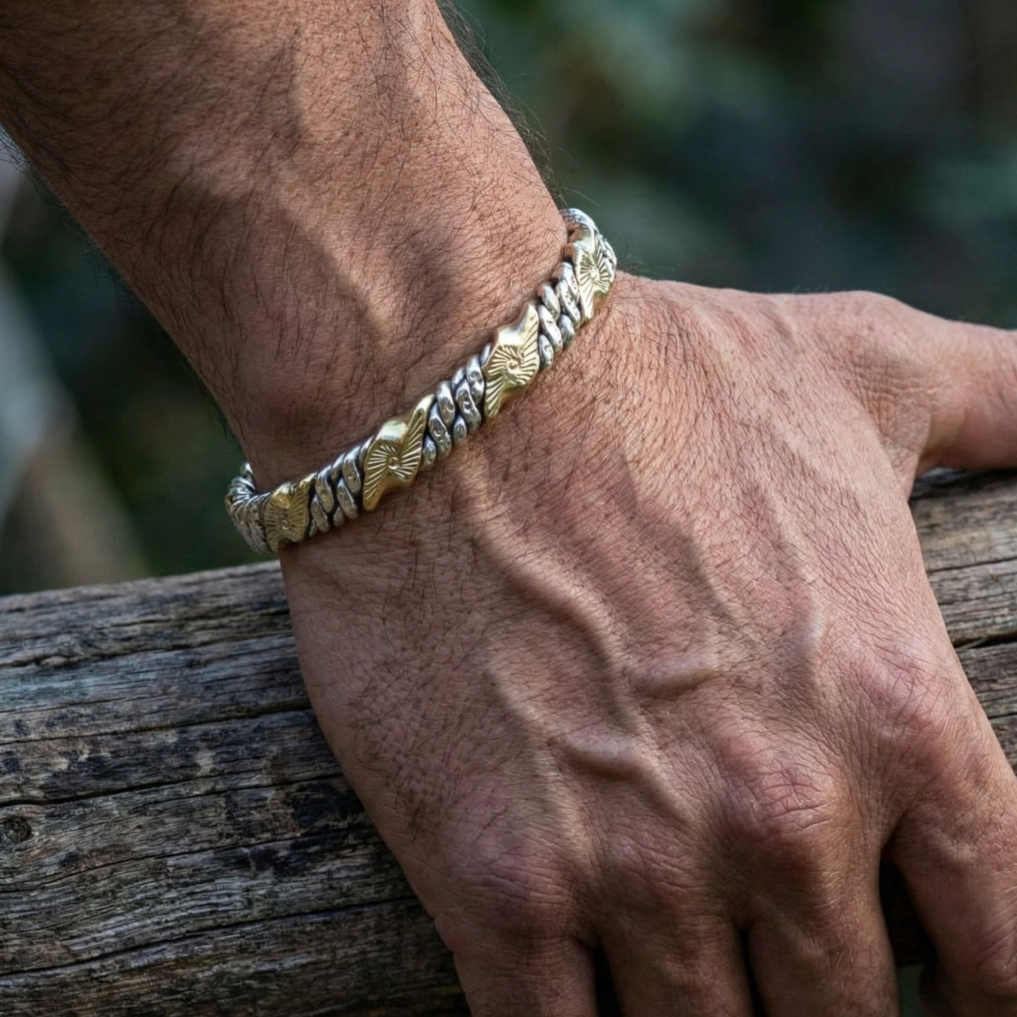 A close-up of a man's rugged hand firmly gripping a weathered wooden log. He is wearing a twisted two-tone metal cuff bracelet. The lighting highlights the texture of his skin and the detailed engraving on the bracelet's brass sections.