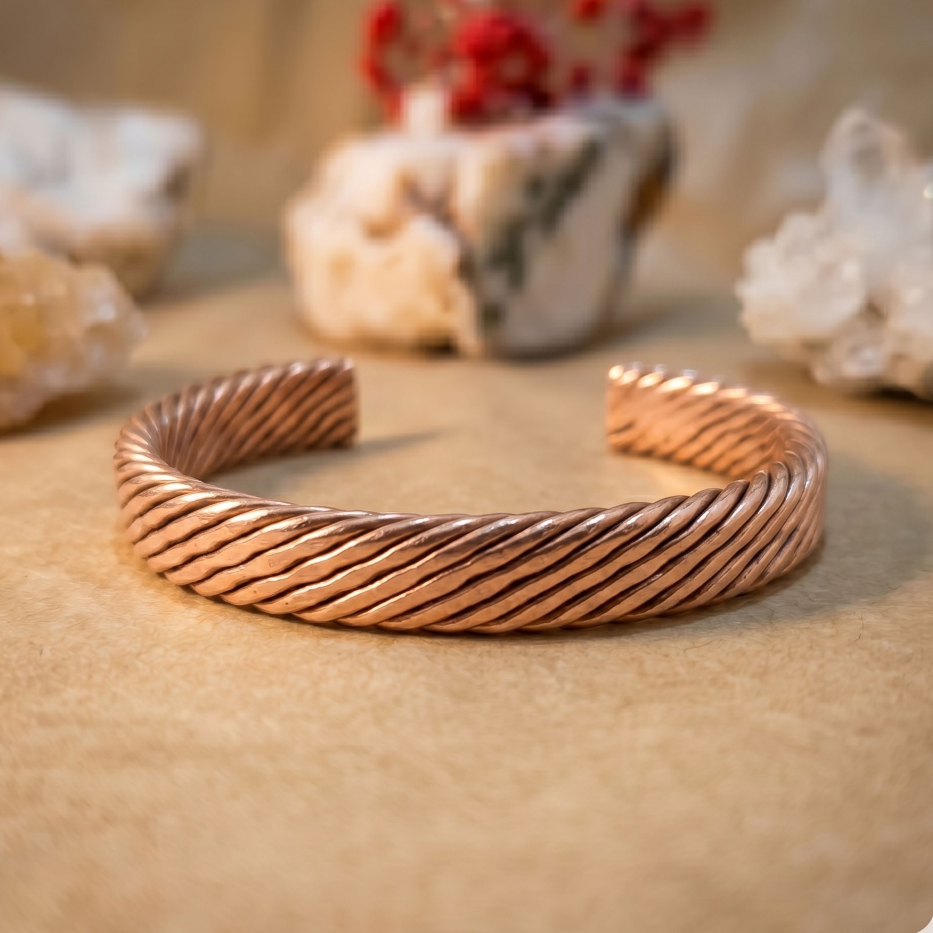A close-up of a finely woven pure copper bracelet resting on an earthy surface, accompanied by raw white crystal clusters and dried red flowers in the softly blurred background
