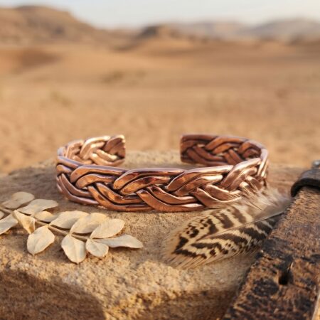 A rustic side-view photograph of the heavy braided pure copper men's bracelet resting on a rough, natural desert sandstone rock. The eye-level angle emphasizes the height and density of the handwoven pattern