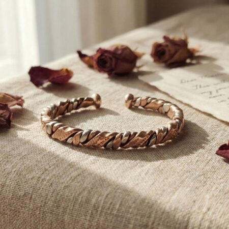 A twisted copper and silver-tone cuff bracelet resting on soft beige linen, surrounded by dried rosebuds and a vintage handwritten note, creating a romantic aesthetic.