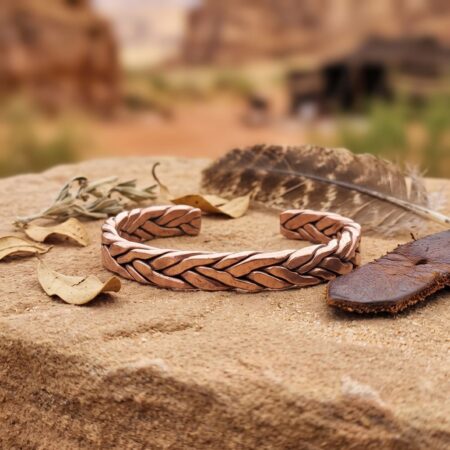 A rustic, heavy woven pure copper men's bracelet resting on a flat sandstone rock outdoors, decorated with dried leaves and a bird feather.