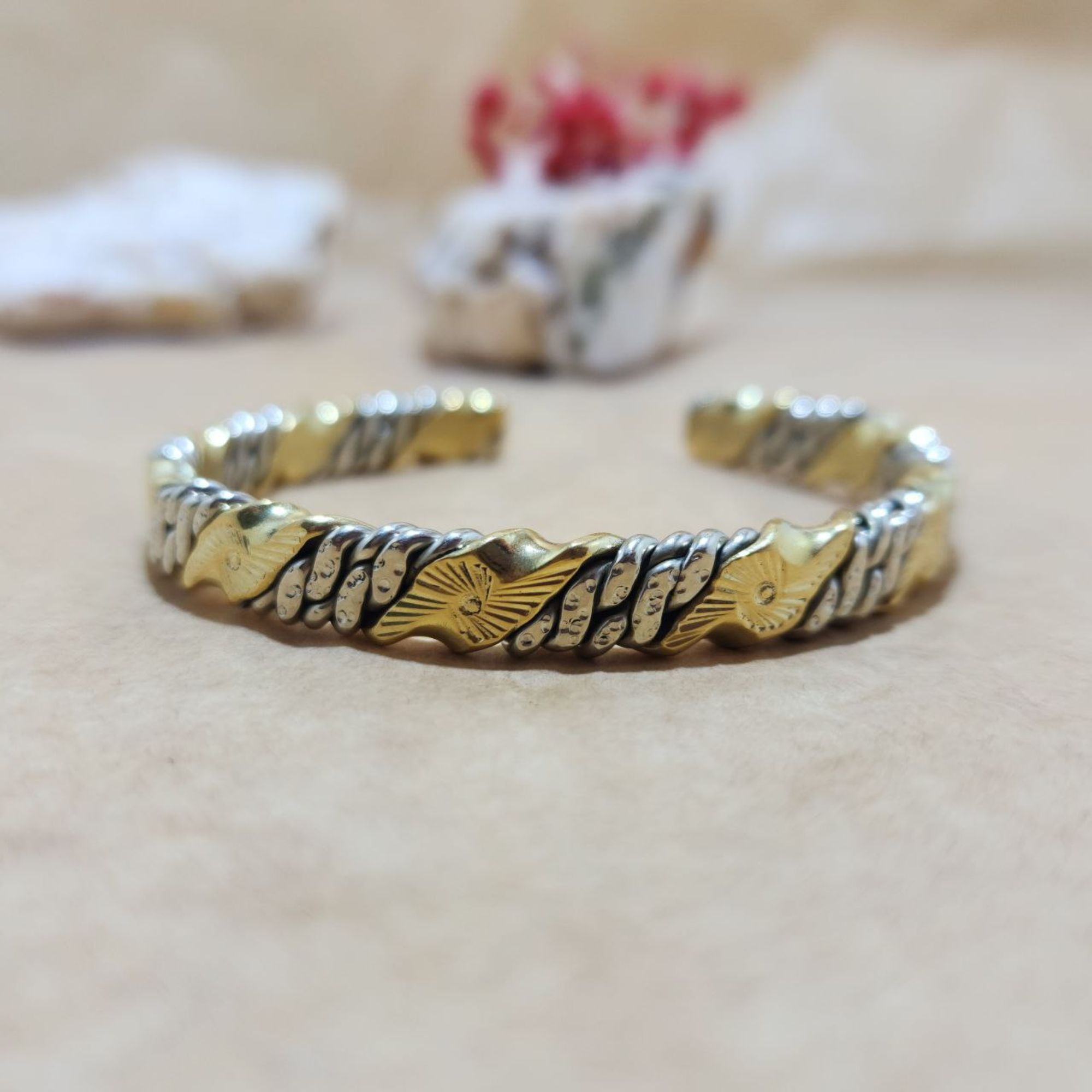 A close-up studio shot of a mixed-metal twisted cuff bracelet made of stainless steel and brass. It is resting on a beige surface with blurred white stones and a small red dried flower arrangement in the background.