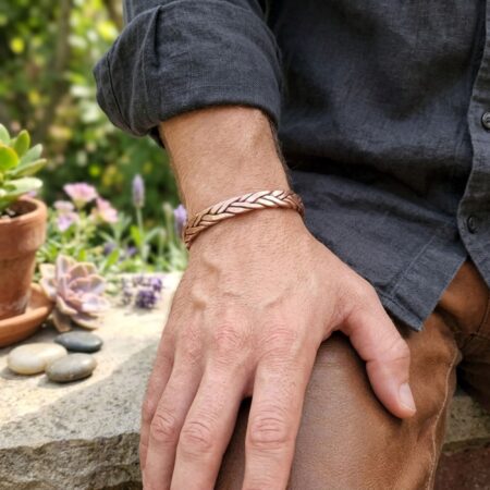 A lifestyle shot of a man wearing a dark linen shirt and a braided copper strength bracelet on his wrist, resting his hand casually in an outdoor garden setting.