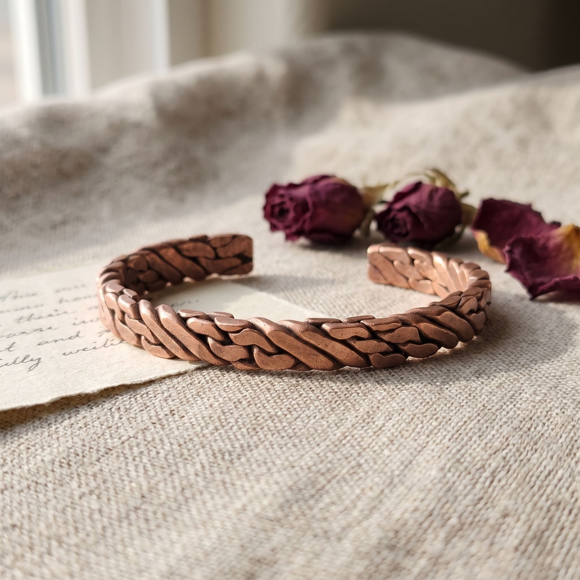 A thick, hand-forged pure copper woven men's bracelet resting on a piece of vintage script paper and textured linen, accented in the background by dried red roses.