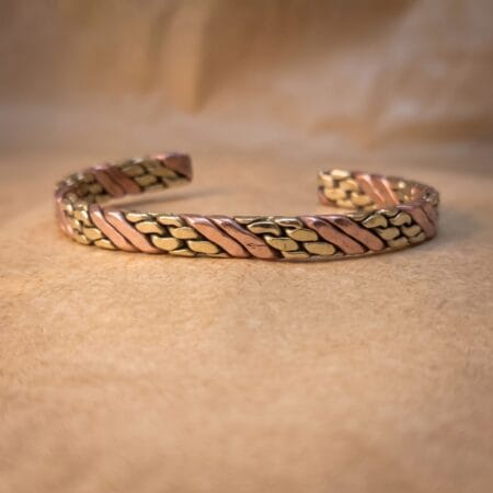 A macro studio shot of a hand-forged brass and copper interlocking woven men's cuff resting on a warm, earth-toned surface to emphasize the contrasting raw metals.