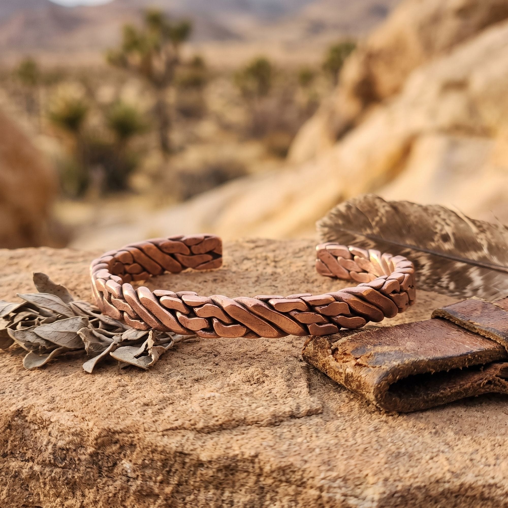 A rugged, pure copper men's viking bracelet resting on a rough stone outdoors, styled next to a weathered leather strap and a bird feather, with a blurred desert landscape in the background