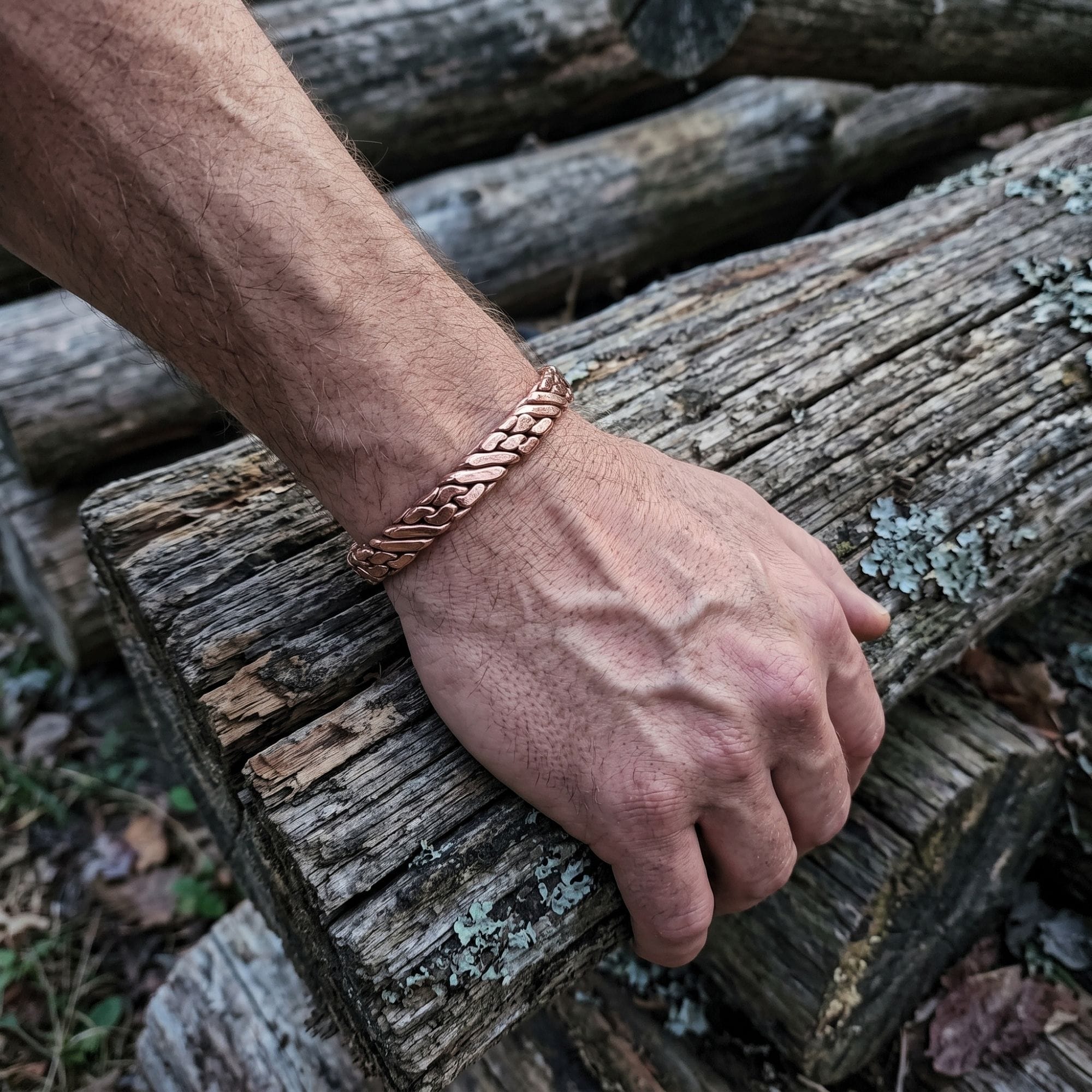 A man's arm wearing a heavy woven pure copper cuff bracelet, resting against a stack of rough, textured tree logs outdoors to highlight its brutalist style