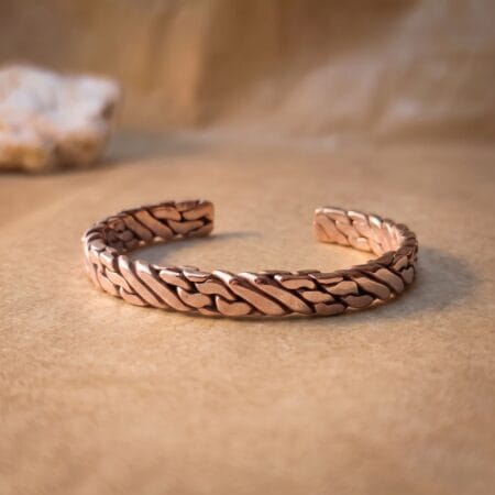A thick, polished pure copper hand-woven men's cuff bracelet resting on a smooth, warm-toned surface with soft studio lighting emphasizing the interlocking metal strands.