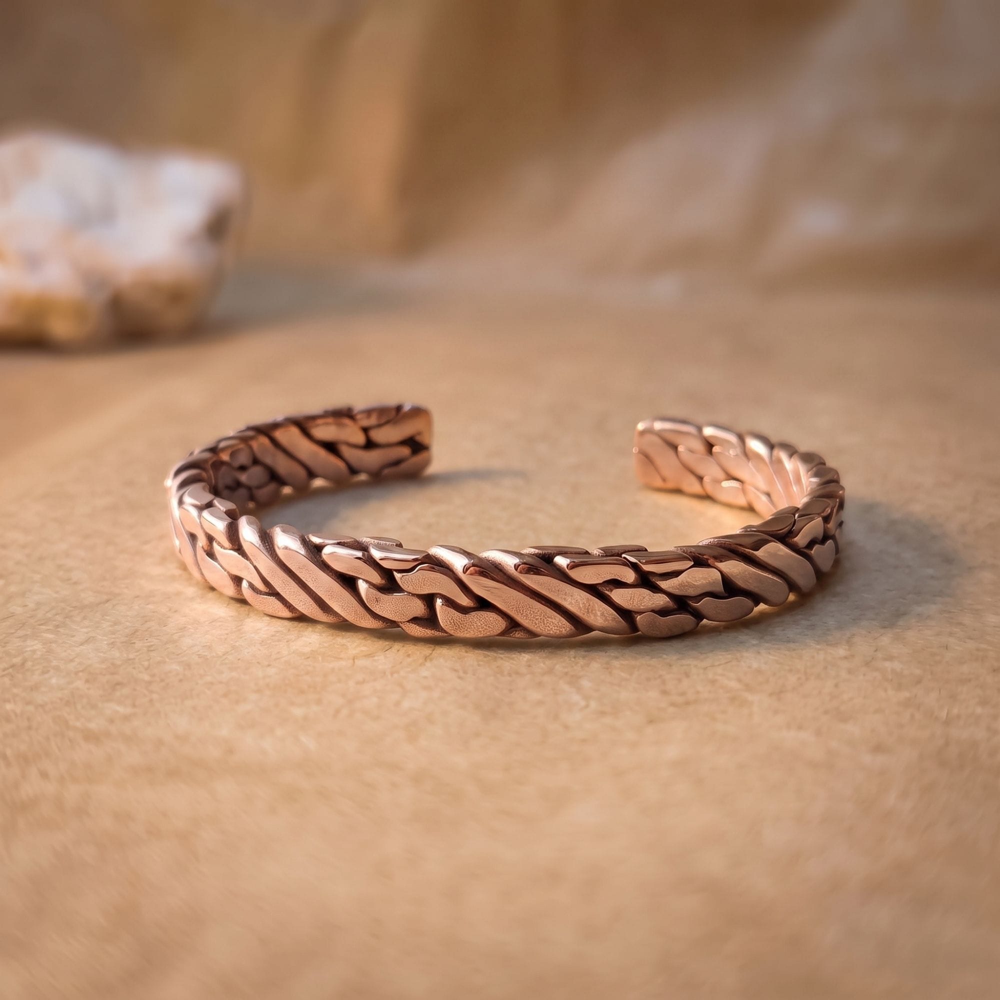 A thick, polished pure copper hand-woven men's cuff bracelet resting on a smooth, warm-toned surface with soft studio lighting emphasizing the interlocking metal strands.
