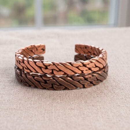 Two heavy woven men's viking bracelets stacked together on a light linen fabric. The top cuff features bright, raw solid copper, while the bottom cuff features a dark, oxidized copper finish.